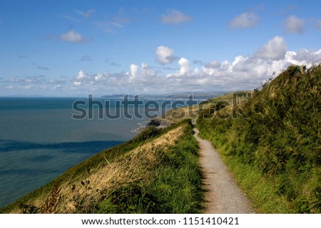 Sunny, summer day view along narrow grass lined cliff top path way with deep blue sea and cliff view and mountains and countryside on the horizon, Ceredigion Coastal Path, Aberystwyth and Clarach Royalty-Free Stock Photo #1151410421