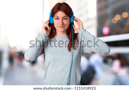 Young redhead girl listening to music with headphones on unfocused background