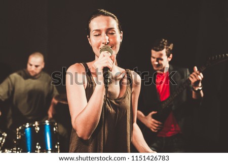 Closeup of a young woman singing with passion while the drummer and the guitarist are in the blurry background.