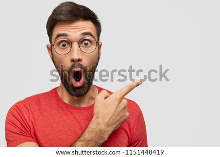 Surprised bearded young male with eyes popped out, has astonished expression, points aside at blank copy space for your advertisement or promotional text. People, emotions, reaction concept. Royalty-Free Stock Photo #1151448419