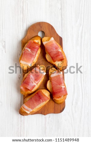 Crostini with serrano ham on rustic wooden board over white wooden background, top view.