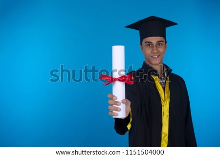 Young smiley student wear graduation robe and cap holding his rolled certificate showing it to the camera on a blue background.