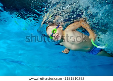 A beautiful little boy is swimming under the water in the pool on a blue background, looking forward and smiling. Portrait. Horizontal orientation.