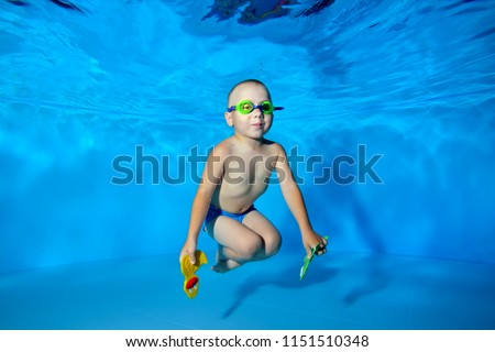 A happy little boy swims and poses underwater in the pool on a blue background with swimming glasses and toys in his hands. He looks at the camera and smiles. Portrait. Underwater photography.
