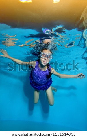 Funny little girl is swimming underwater in the pool on yellow light background in swimming glasses and purple dress. Portrait. Underwater photography. Vertical view.