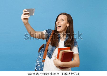 Young happy amazed woman student with opened mouth with backpack holding school books doing taking selfie shot on mobile phone isolated on blue background. Education in high school university college