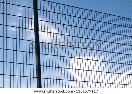 Close-up of high abstract geometrical protective dark metal fence of soccer or football field playground on clear blue sky with bright white clouds background.