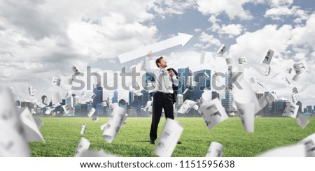 Young and confident businessman in suit starting launching huge white arrow to the air while standing among flying papers on green lawn and cityscape view on background.