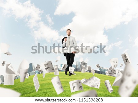 Confident and young businessman in suit holding big white arrow in hands which pointing to the side while standing among flying papers on green lawn and cityscape view on background.