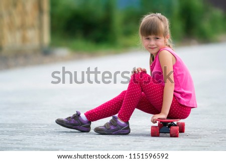 Pretty young long-haired blond child girl in casual pink clothing sitting on skateboard on paved suburb street on blurred sunny summer green background. Children activities, games and fun concept.