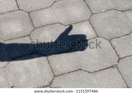 Close-up shot of shadow silhouette of human arm and hand with thumb-up success gesture on gray pavement lit by bright sun.