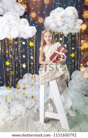 Magic christmas card with a little girl in a studio with a gold stars garland, fluffy clouds and lights. Cute girl in a gold dress sits on a white ladder