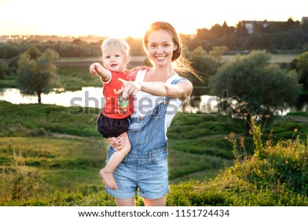 A young, beautiful mother holds a baby boy in her arms and smiles. Mom and son show a handshake in one direction standing in a park outdoors at sunset