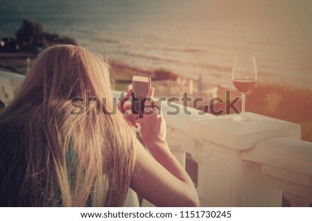 Blonde young woman with long natural hair standing on balcony near the sea and using mobile phone to take photo