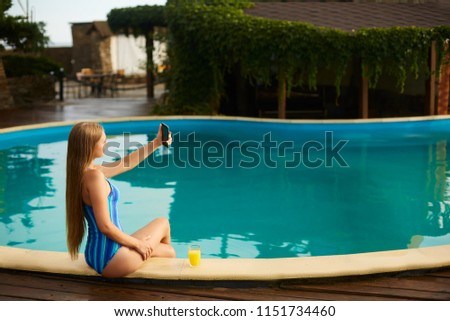 Attractive blonde woman with long hair sitting at poolside and taking selfie portrait with cellphone camera. Slim girl wear blue striped body swimsuit and holds a glass with orange juice.
