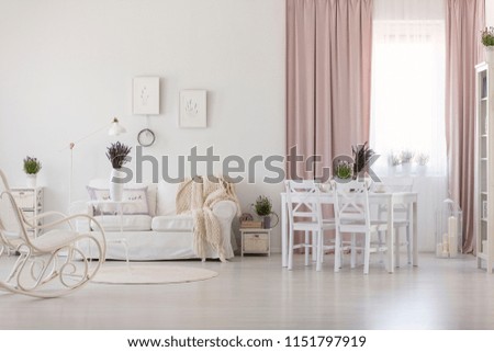 White chairs at dining table in apartment interior with blanket on sofa and pink drapes. Real photo