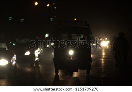 Silhouetted New Dehli traffic at night. Center of the image is an Indian motorcycle taxi, cars on the left of the image and a cyclist on the right.