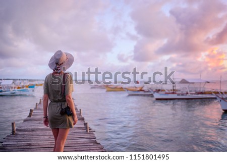 Photography and travel. Young woman in hat holding camera standing on wooden fishing pier with beautiful tropical sea view.