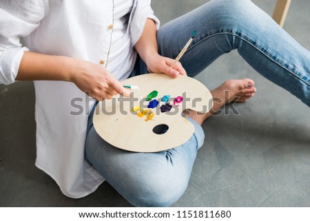 Crop of girl in white shirt and jeans, sitting at work place and creating, drawing picture with oil colorful paints. Pretty woman, female artist holding brush and palette. Concept of art.