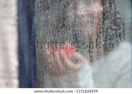 Water Drops on a window glass in a rainy day with a woman holding a red hot coffee inside background, lifestyle concept in the lonely day 