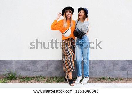 Full height image of two happy cheeky girls , best friends having fun , laughing on white  wall .  Outdoor. Autumn outfit 