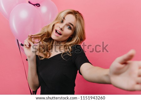 Blissful birthday girl making selfie on pink background. Blonde curly woman taking picture of herself with party balloons and expressing amazement.