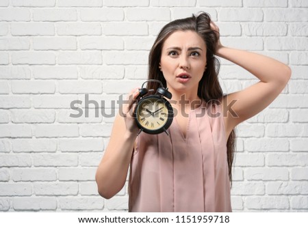 Troubled woman with alarm clock against white brick wall. Time management concept
