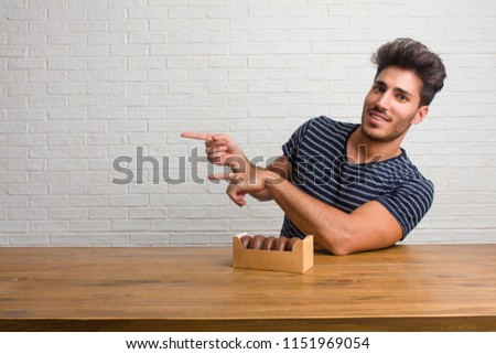 Young handsome and natural man sitting on a table pointing to the side, smiling surprised presenting something, natural and casual. Eating chocolate doughnuts.
