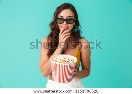 Photo of beautiful brunette woman 20s wearing glasses holding bucket with popcorn and looking at camera isolated over blue background