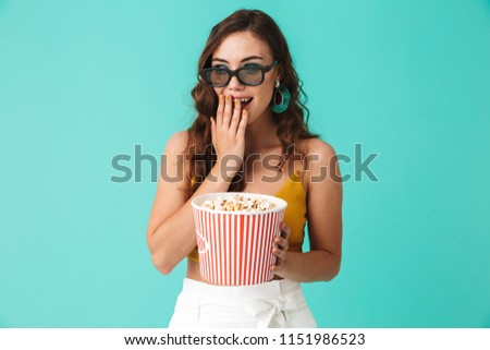 Photo of surprised brunette woman 20s wearing glasses holding bucket with popcorn and looking at camera isolated over blue background