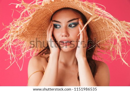 Image closeup of trendy fashionable woman 20s wearing big straw hat holding face and looking aside isolated over pink background