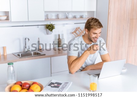 smiing young man drinking coffee and looking at laptop screen at kitchen  Royalty-Free Stock Photo #1151998517