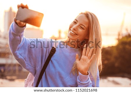 Portrait of a pretty young girl with backpack standing outdoors during sunset, taking selfie with mobile phone, waving