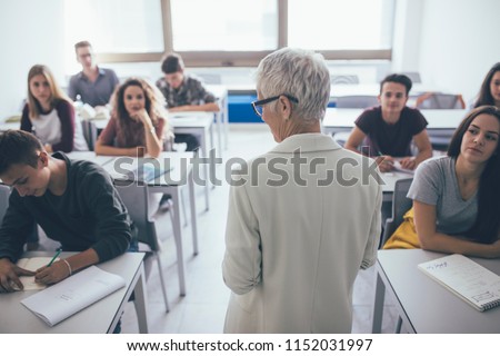 Back view of grey-haired woman teacher stading at classroom and talking to high school students. Royalty-Free Stock Photo #1152031997