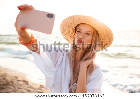 Pretty young girl in summer hat and swimwear spending time at the beach, taking a selfie with outsretched hand