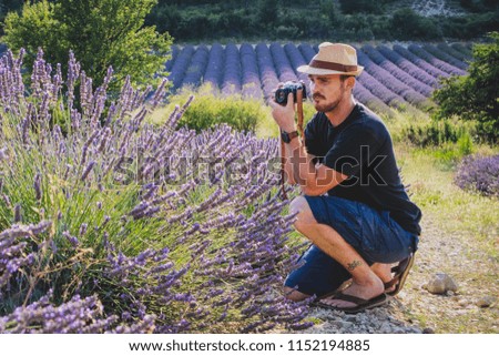 A young caucasian male with hipster image and thatched hat is taking a picture of a blooming lavender field with a vintage photo camera. Person looking away from camera.