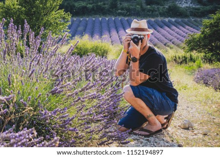 A young caucasian male with hipster image and thatched hat is taking a picture of a blooming lavender field with a vintage photo camera. Person looking towards the camera.