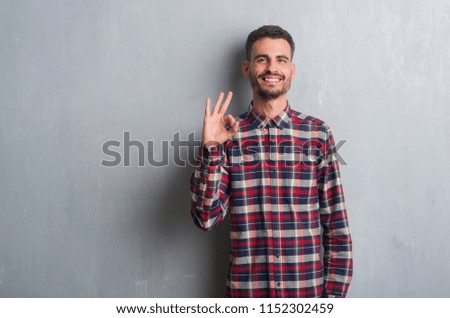 Young adult man standing over grey grunge wall doing ok sign with fingers, excellent symbol