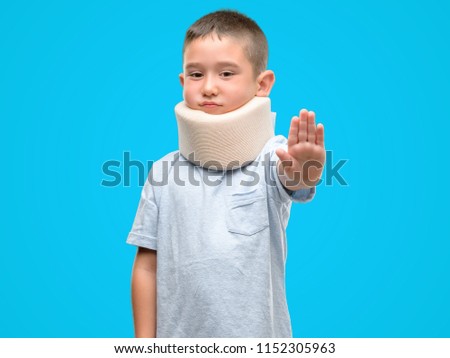 Dark haired little child injured wearing neck collar with open hand doing stop sign with serious and confident expression, defense gesture