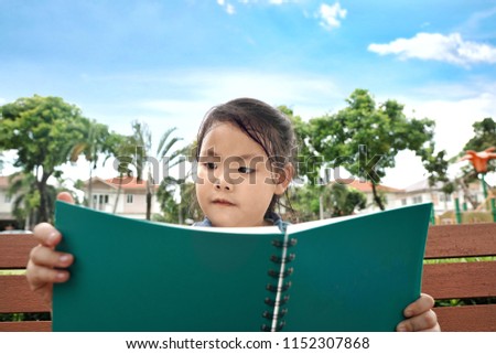 Asian little cute girl kid student in uniform school reading a book sitting on the wooden bench in the park with cloud and blue sky background.Education study learning and success literacy concept.