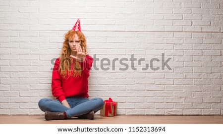 Young redhead woman sitting over brick wall wearing birthday hat with open hand doing stop sign with serious and confident expression, defense gesture