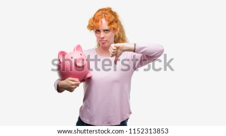 Young redhead woman holding piggy bank with angry face, negative sign showing dislike with thumbs down, rejection concept