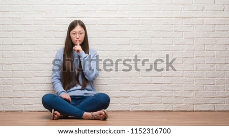 Young Chinese woman sitting on the floor over brick wall asking to be quiet with finger on lips. Silence and secret concept.