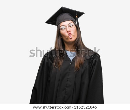 Young hispanic woman wearing graduated cap and uniform making fish face with lips, crazy and comical gesture. Funny expression.