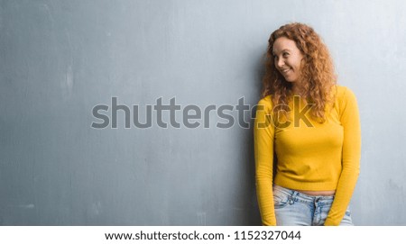 Young redhead woman over grey grunge wall looking away to side with smile on face, natural expression. Laughing confident.