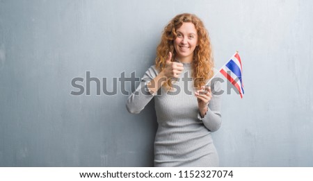Young redhead woman over grey grunge wall holding flag of Thailand happy with big smile doing ok sign, thumb up with fingers, excellent sign