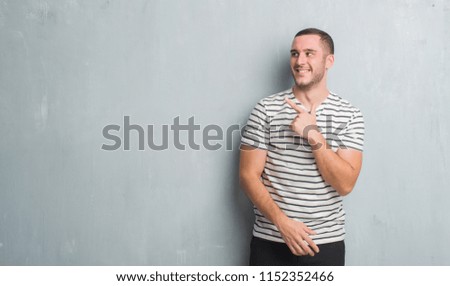 Young caucasian man over grey grunge wall cheerful with a smile of face pointing with hand and finger up to the side with happy and natural expression on face looking at the camera.