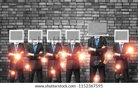Businessmen in suits with monitors instead of their heads keeping arms crossed while standing in a row and one at the head with laptop in empty room against gray wall on background.
