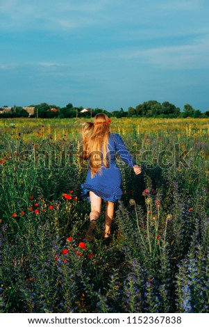 Girl with long blond hair wearing blue retro polka dot dress looking like hippie walking outdoors in the field of wild flowers