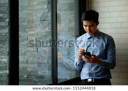 Confident young Asian businessman working on digital tablet while standing in side of the big window in office 